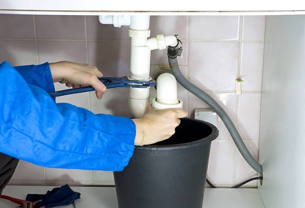 A plumber using an adjustable wrench to fix a sink's piping system, with a bucket underneath to catch drips.