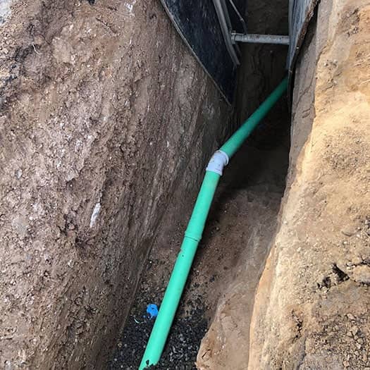 A green pvc pipe installed in a narrow trench with exposed soil walls, leading to a wooden framed structure.