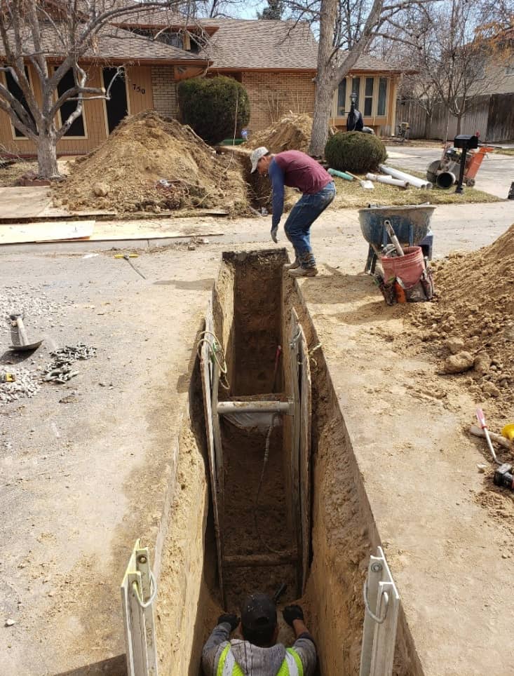 Two workers excavate and reinforce a deep trench in a residential area, with equipment, soil piles, and a wheelbarrow nearby.