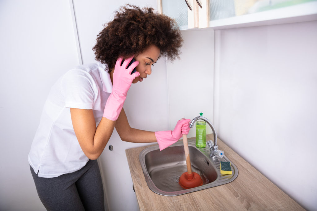 Close-up of a woman calling plumber on mobile phone while cleaning the sink with plunger.