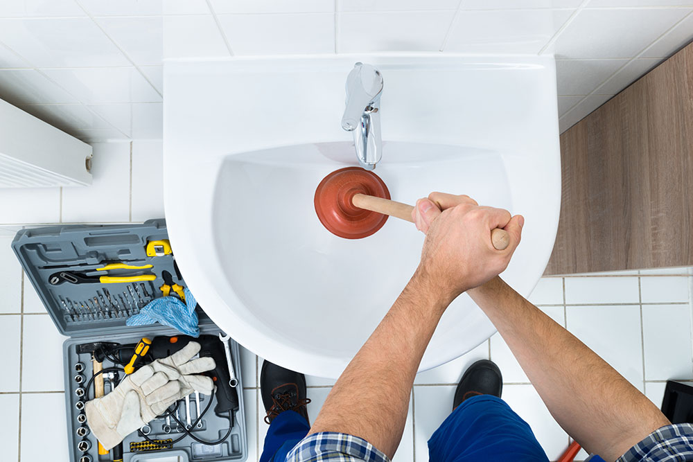 A man has tools on the floor and is trying to plunge a clogged bathroom sink