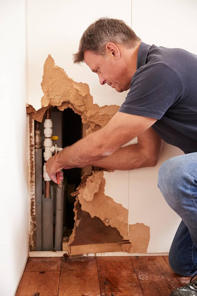 A plumber is doing an emergency repair to a burst pipe in an interior wall behind drywall