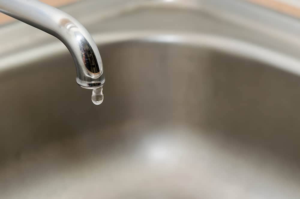 A kitchen sink faucet is leaking and has a drop of water falling from it