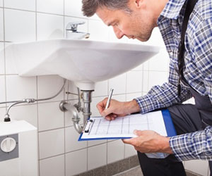A plumber is diagnosing a bathroom sink drain problem with a clipboard in his hands.