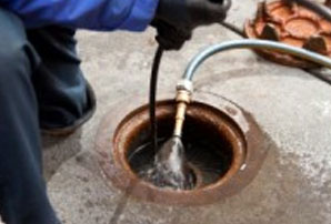 A plumber is accessing an outdoor sewer drain and cleaning it with a drain auger and water.