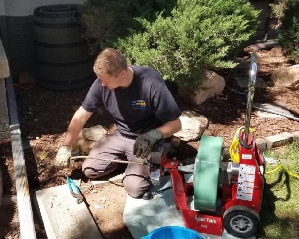 A person wearing gloves operates plumbing equipment and works on an outdoor pipe near bushes and a house.