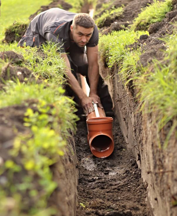 A man installs a section of orange PVC drainage pipe in a narrow, muddy trench lined with grass and soil.