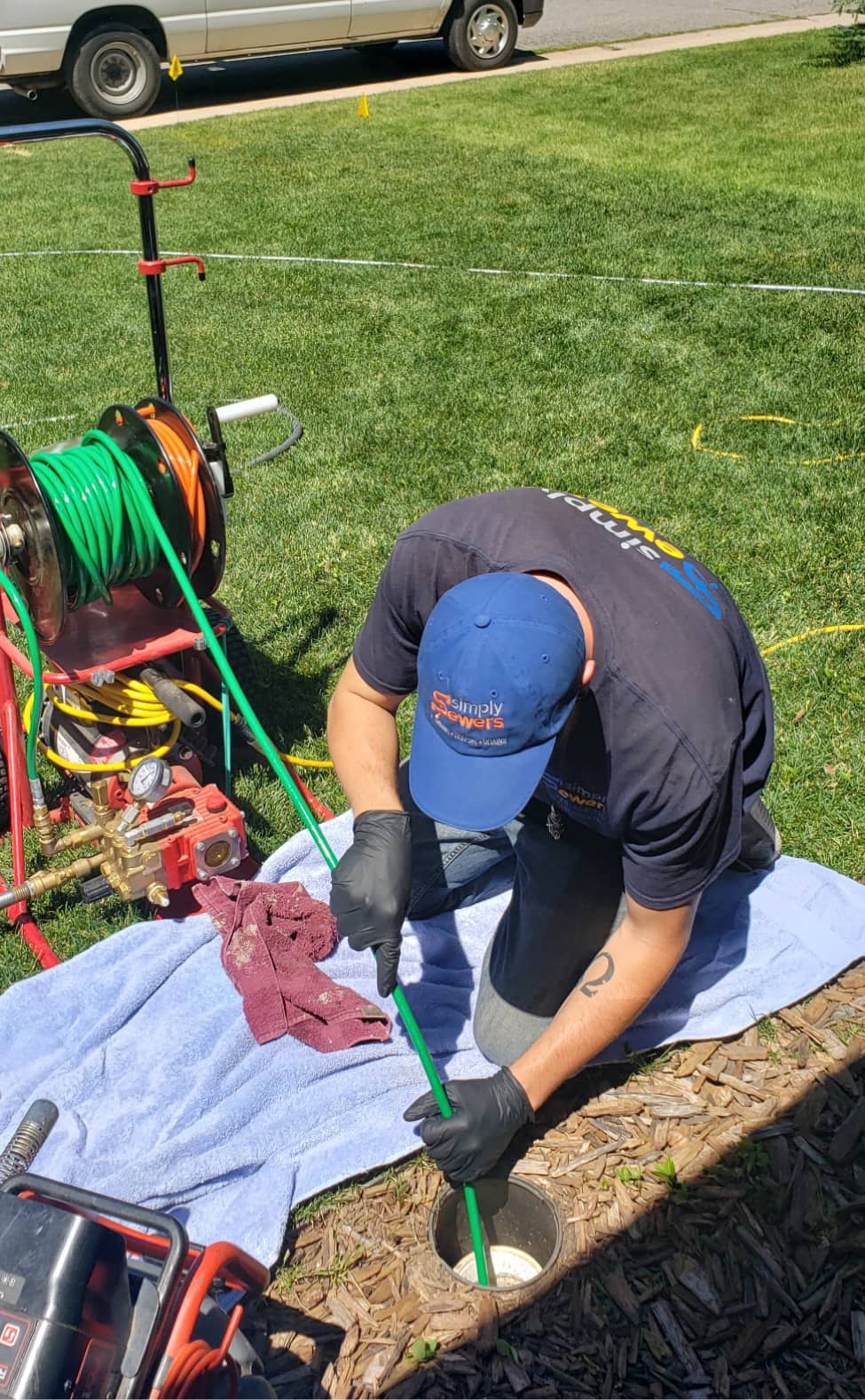 A worker wearing gloves and a hat kneels on a towel while using a green hose to clean or inspect an outdoor pipe opening on a grassy lawn.