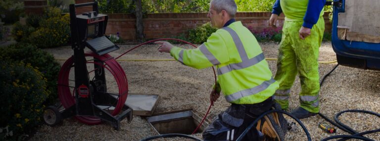 Two workers in high-visibility clothing use equipment to inspect or clean a drain or pipe on a gravel driveway.