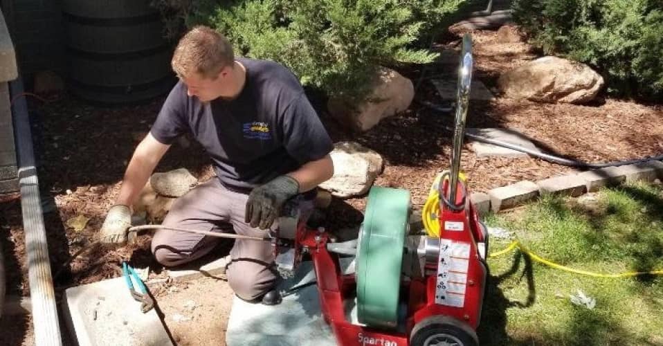 A man kneels on the ground outdoors using a drain cleaning machine with hoses and gloves, surrounded by garden plants and landscaping rocks.