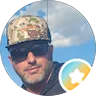 Man wearing a floral cap and sunglasses, standing outdoors with a partly cloudy sky in the background. A round badge with a star is in the lower right corner.