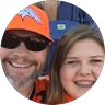 Smiling man in a Denver Broncos hat and sunglasses poses for a selfie with a young woman at a sports stadium.