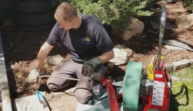 A person wearing gloves uses a plumbing auger machine outdoors, feeding a cable into a pipe surrounded by rocks and plants.