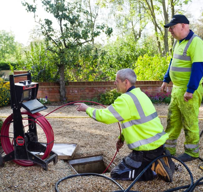 Two workers in high-visibility clothing operate equipment and inspect cables near an open utility access point in a garden area.